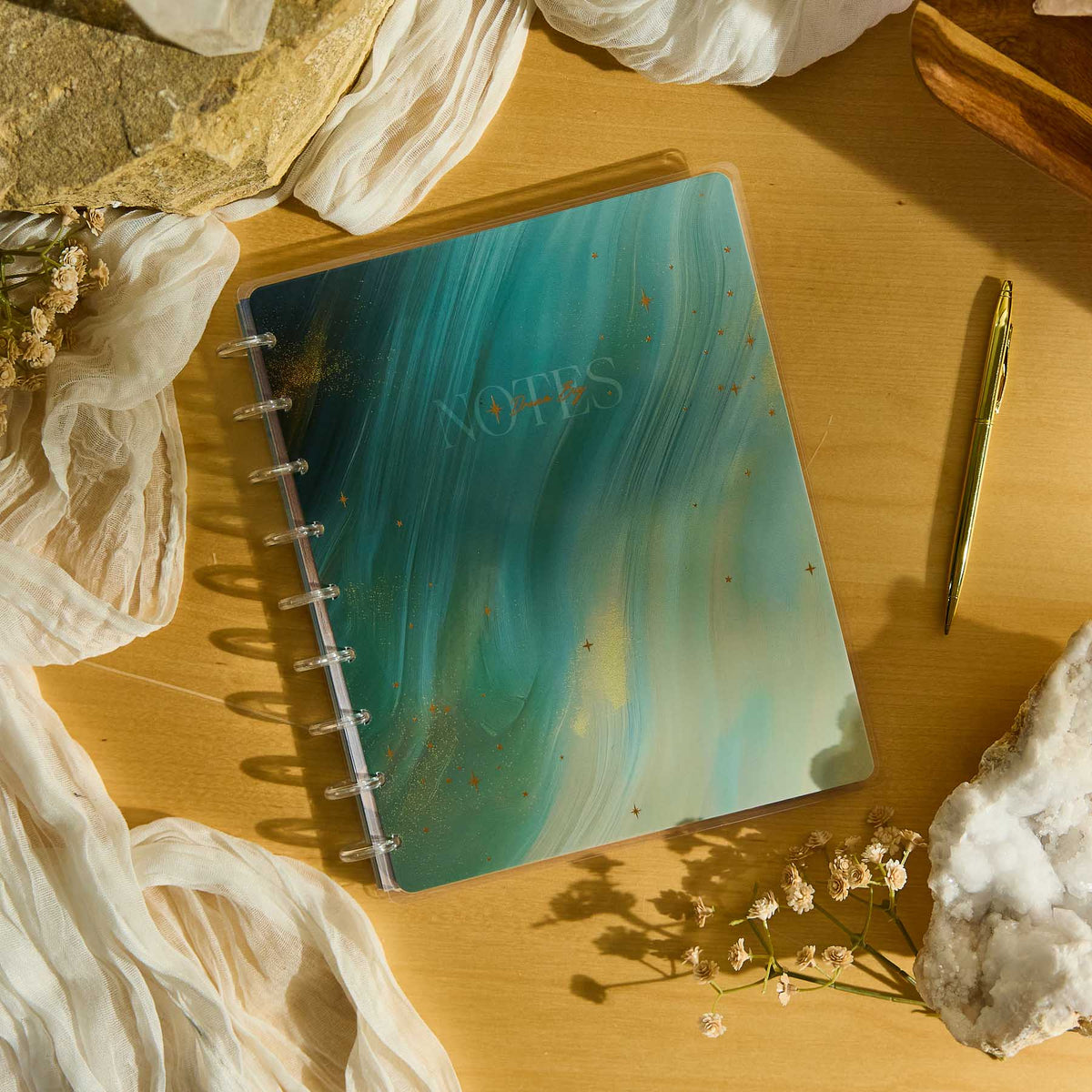Starlit Glow notebook with a teal marbled cover beside a gold pen and dried flowers