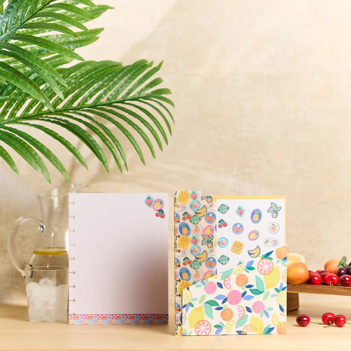 A decorative notebook or journal set displayed on a tabletop with a palm leaf, glass pitcher, and fruit bowl in the background. The set includes a white page-covered notebook with a colorful patterned spine and matching patterned cover, and a smaller matching notebook standing beside it.