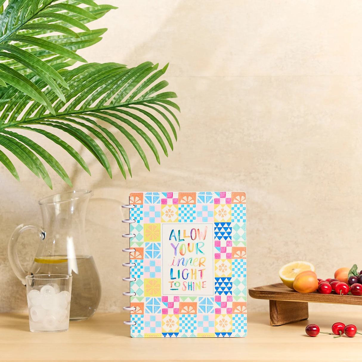A colorful spiral-bound notebook with a patchwork pattern and the text 'ALLOW YOUR INNER LIGHT TO SHINE' sits on a wooden table next to a glass pitcher, a green plant leaf, and a small fruit stand.
