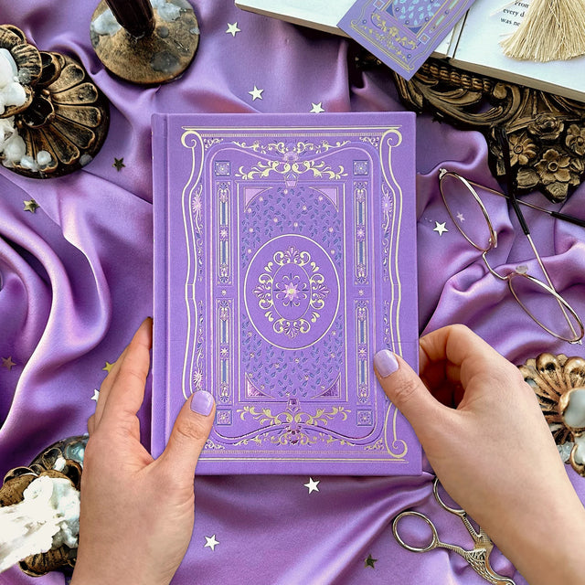 Literati Wisteria Journal Decorative purple book held by hands on a purple fabric background with gold accents.