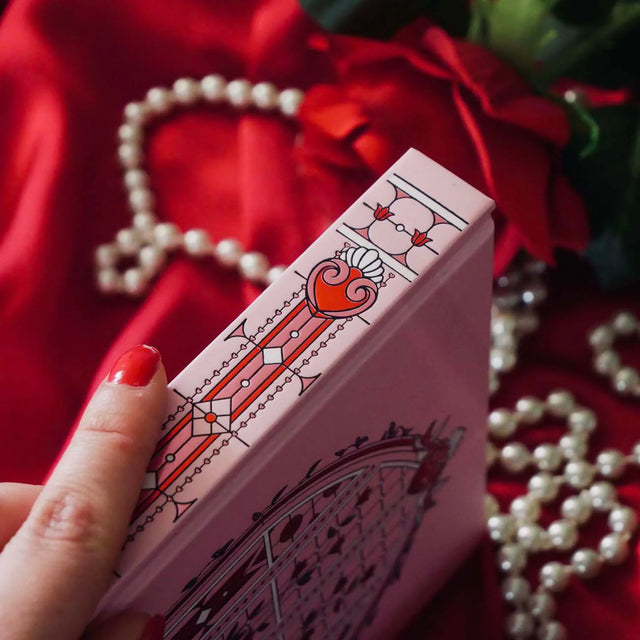 Pink notebook with decorative design held by a hand against a red fabric and pearl background