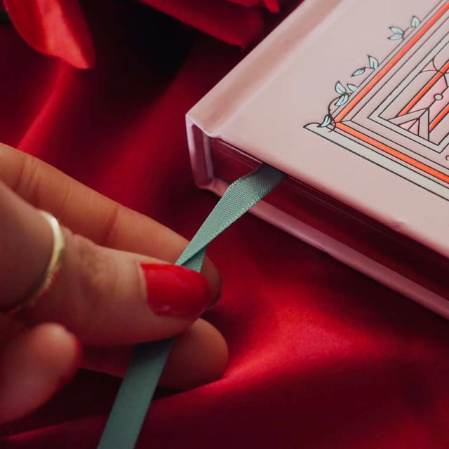 Hand with red nail polish holding a blue ribbon on a pink book against a red fabric background