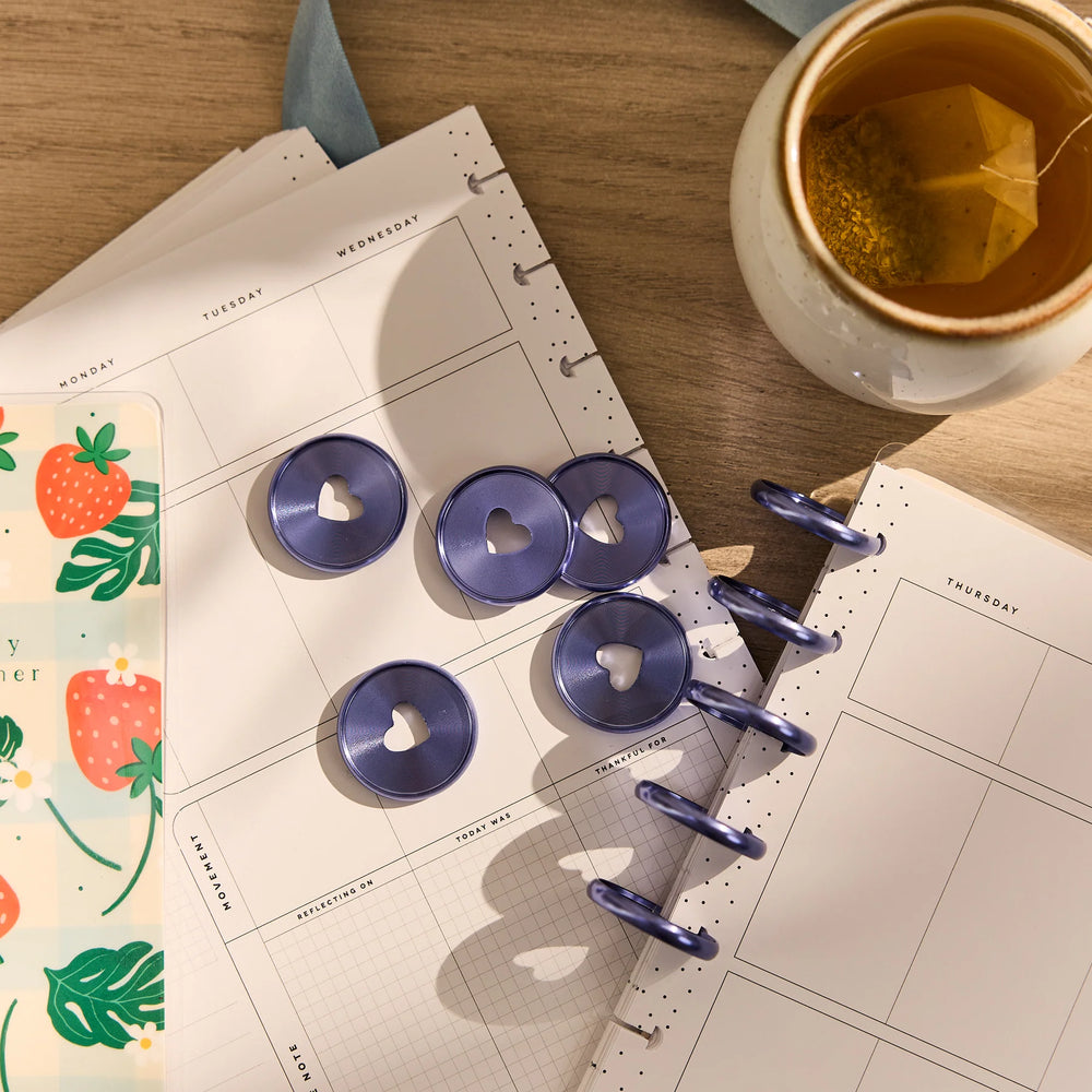 Purple heart-shaped planner clips on a open planner with a cup of tea and colorful card.