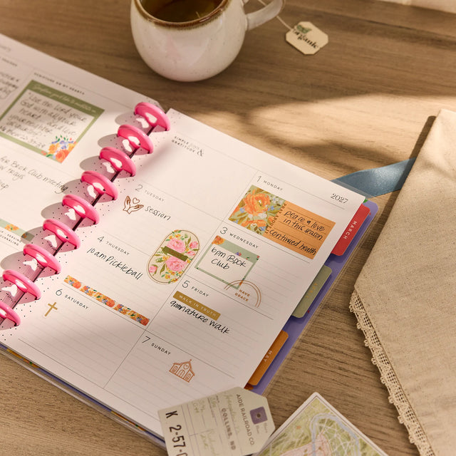 Open planner with colorful stickers on a wooden surface next to a cup of tea.