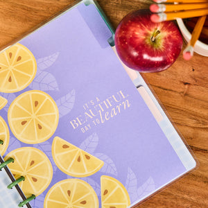 Notebook with lemon design and text 'It's a beautiful day to learn', apple, and pencils on a wooden surface