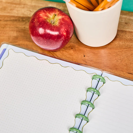 Red apple next to a cup of carrots on a wooden table with a notebook in the foreground.