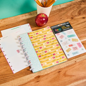 Happy Planner Pawsitive Notebook + Sticker Bundle, notebook colorful cover, apple, and Happy Planner stickers on a wooden surface.