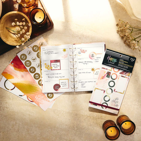 An open disc-bound planner sits on a warm beige surface with decorative cards, stickers, and a packaged stamp set around it. The scene includes candles, dried flowers, and a wooden tray in a styled flat-lay arrangement.