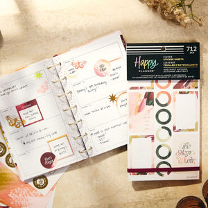 An open ring-bound planner with decorated weekly pages lies next to a package of Happy Planner little binder sheets. The scene is styled on a warm beige textured surface with dried flowers.