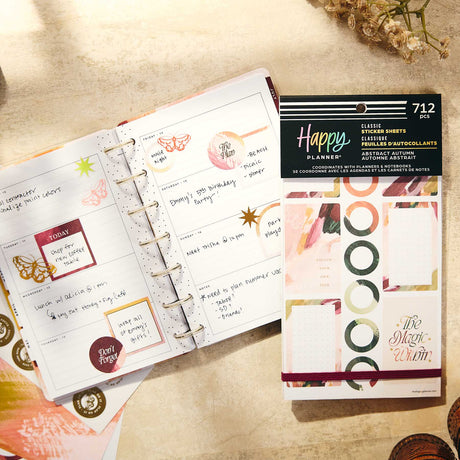 An open ring-bound planner with decorated weekly pages lies next to a package of Happy Planner little binder sheets. The scene is styled on a warm beige textured surface with dried flowers.