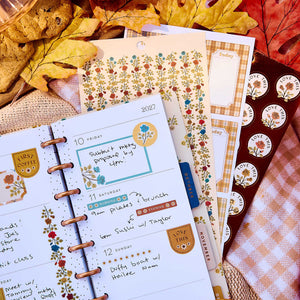 Close-up of a weekly planner decorated with Fall Picnic boxes and florals beside sticker sheets and autumn leaves