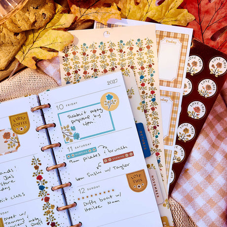 Close-up of a weekly planner decorated with Fall Picnic boxes and florals beside sticker sheets and autumn leaves
