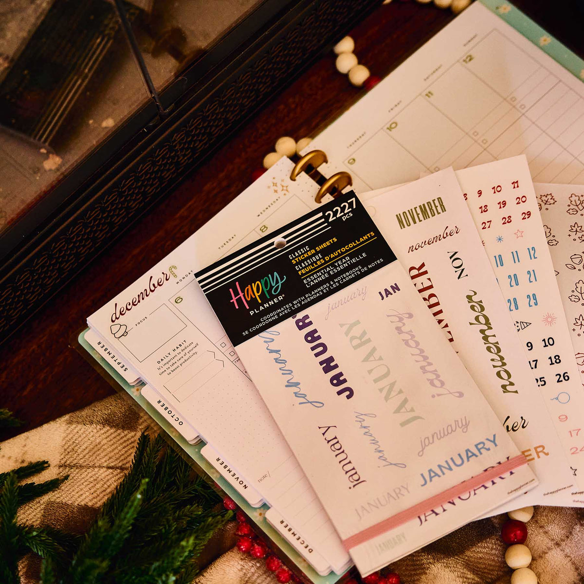 Happy Planner Essential Year sticker book and loose month stickers on an open planner beside seasonal decorations