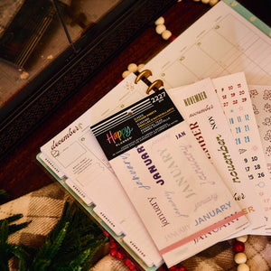 Happy Planner Essential Year sticker book and loose month stickers on an open planner beside seasonal decorations