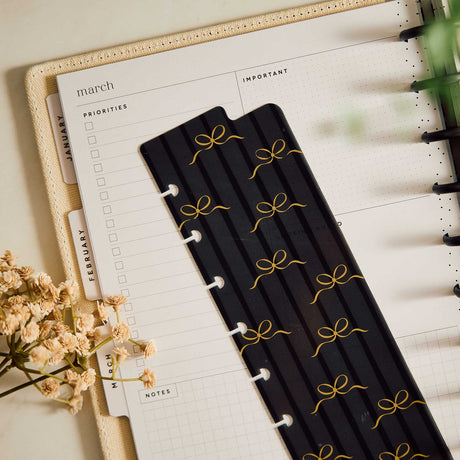 Black Modern Balance bookmark with gold bow detailing resting on an open disc-bound planner