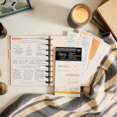 Hushed Hues sticker book and reflection stickers beside an open planner, candle and striped fabric