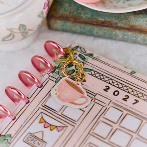 Paper Raven Co. coffee cup charm resting on an illustrated planner beside pink discs