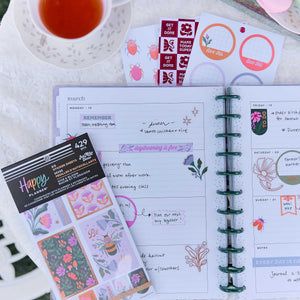 Hive & Bloom floral sticker sheets over an open planner with handwritten plans beside a cup of tea