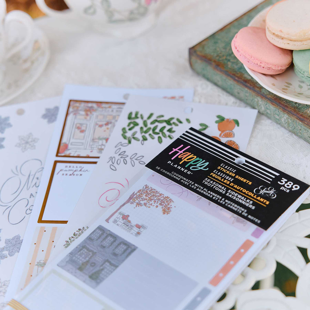 A package of D. La Beige Classic Planner Sheets with autumn-themed designs, fanned out on a white tablecloth next to macarons and a teacup.