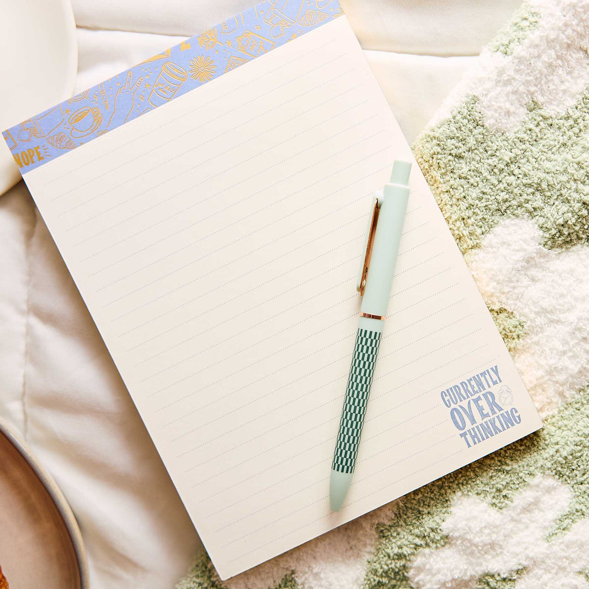 Pajama Plans lined notepad beside a mint green pen on a cream blanket