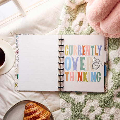 Open Pajama Plans notebook with Currently Overthinking artwork beside coffee and a croissant