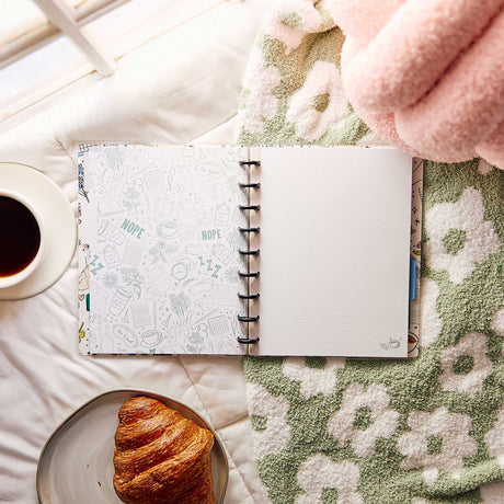 Open Pajama Plans notebook on a blanket beside coffee and a croissant