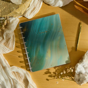 Starlit Glow notebook with a teal marbled cover beside a gold pen and dried flowers