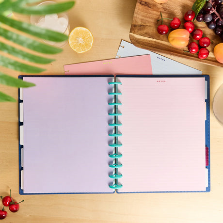 An open spiral-bound planner or notebook with pastel lavender pages and turquoise rings, styled on a light wood tabletop with fruit, a glass of white drink, and green leaves around it.