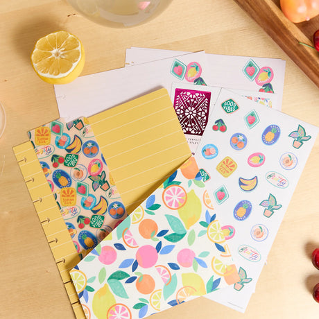 A set of colorful fruit-themed planner stickers and matching decorative paper sheets arranged on a light wood desk, with a lemon half nearby.