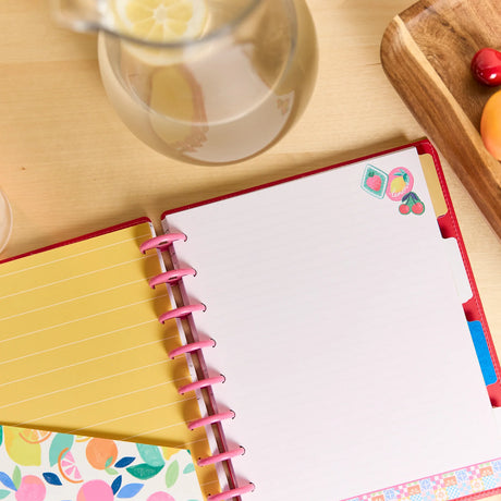 An open spiral-bound planner or notebook with a red cover, yellow lined pages, and colorful fruit-themed stickers on the page, photographed on a light wood tabletop with a glass pitcher and fruit tray nearby.