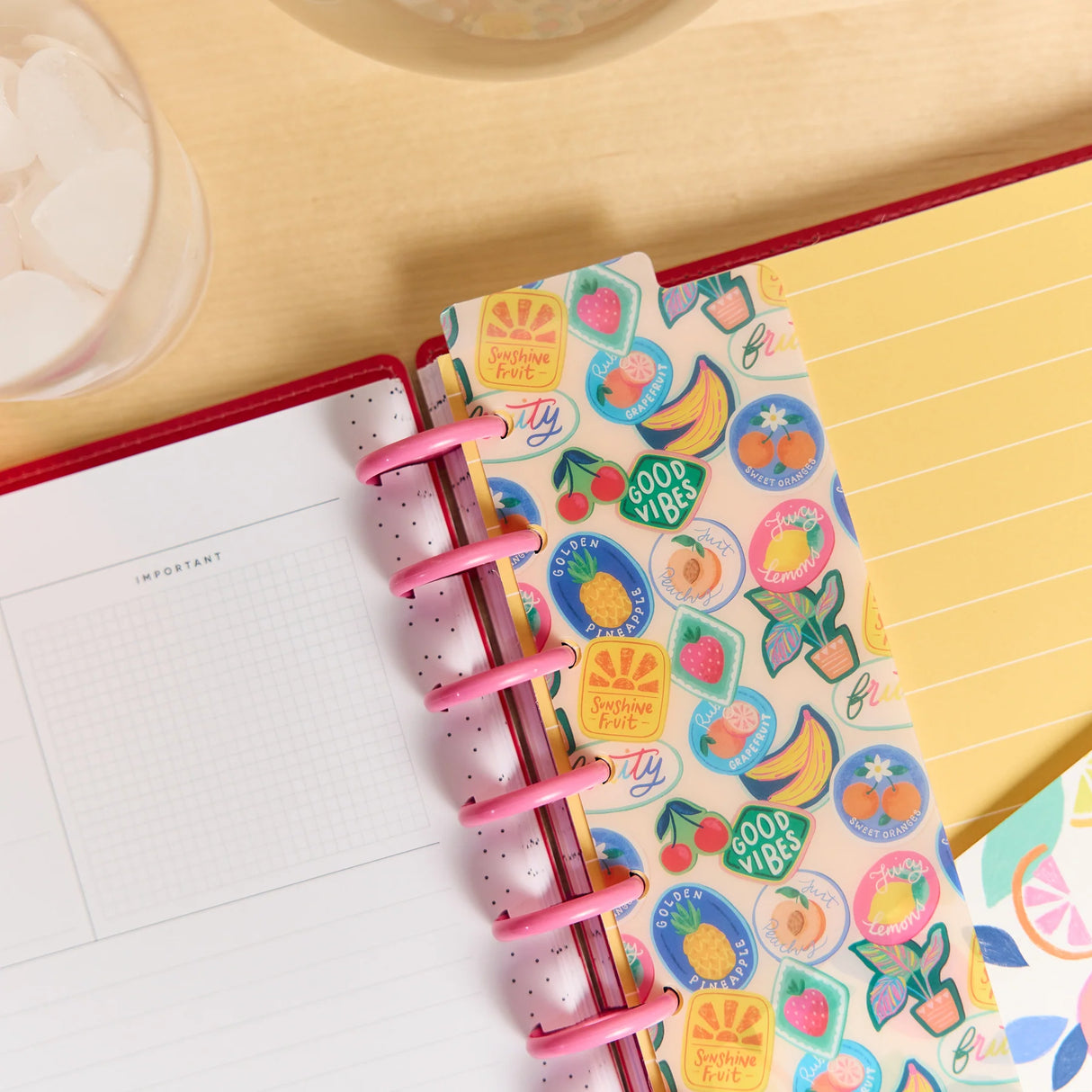 A close-up of a planner or notebook with a colorful fruit-themed sticker strip, pink spiral rings, and a yellow lined page on a light wood desk.