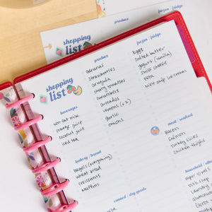 A close-up of an open pink ring-bound planner showing a handwritten shopping list page with columns for produce, beverages, bakery/bread, eggs, meat/vegetables, and household items. A second matching shopping list sheet is partially visible behind it on a light wood surface.