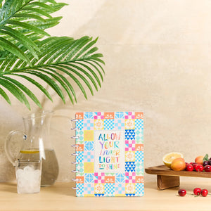 A colorful spiral-bound notebook with a patchwork pattern and the text 'ALLOW YOUR INNER LIGHT TO SHINE' sits on a wooden table next to a glass pitcher, a green plant leaf, and a small fruit stand.
