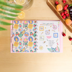Open notebook with decorative stickers on a wooden surface with fruits and a glass of lemonade.