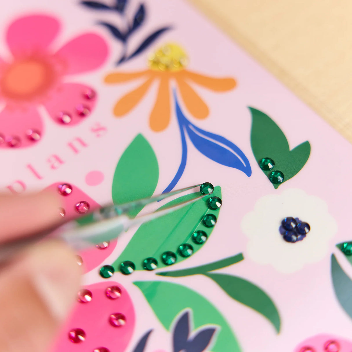 A close-up of a hand using tweezers to place green rhinestones along a decorative green leaf on a pink planner cover decorated with colorful floral illustrations.