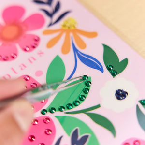 A close-up of a hand using tweezers to place green rhinestones along a decorative green leaf on a pink planner cover decorated with colorful floral illustrations.
