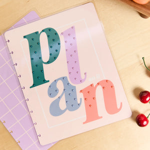 A pastel-colored planner or notebook cover with the word 'plan' spelled out in large decorative letters, photographed on a light wood surface with a few red cherries nearby.