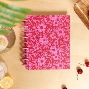 Happy Planner Deluxe Sunshine Splash Classic 12-Month Undated Planner with a pink floral pattern on the cover, styled on a light wood tabletop with decorative props including a glass of lemonade, a lemon slice, cherries, and a wooden board.