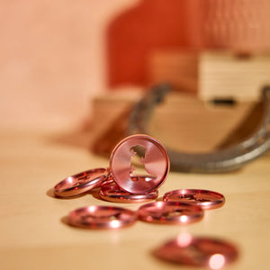 Several metallic rose gold circular buttons with a central cutout, with one standing upright to show the design. The buttons are arranged on a light wooden surface with a warm, blurred background.