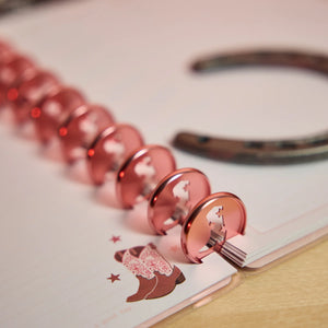 A close-up of a spiral-bound notebook with metallic rose gold binder rings, featuring a cowboy boot and star foil design on the cover, with a blurred horseshoe in the background.