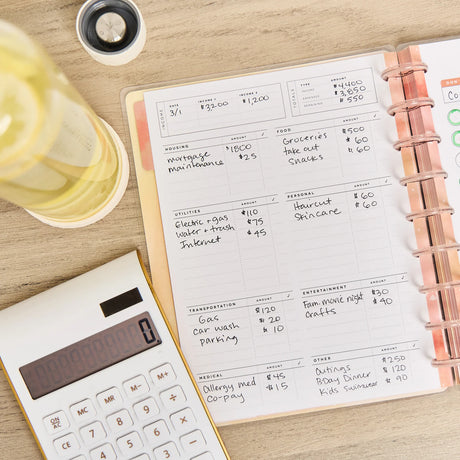 Notebook with budgeting entries on a wooden surface next to a calculator and glass of water.