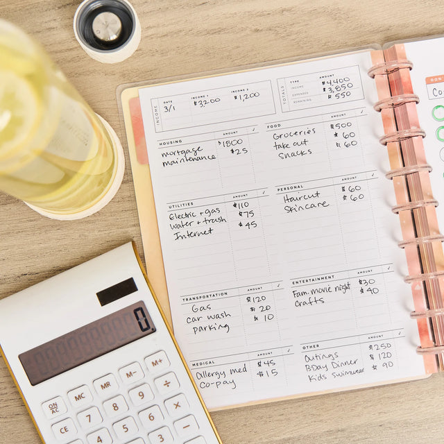 Notebook with budgeting entries on a wooden surface next to a calculator and glass of water.
