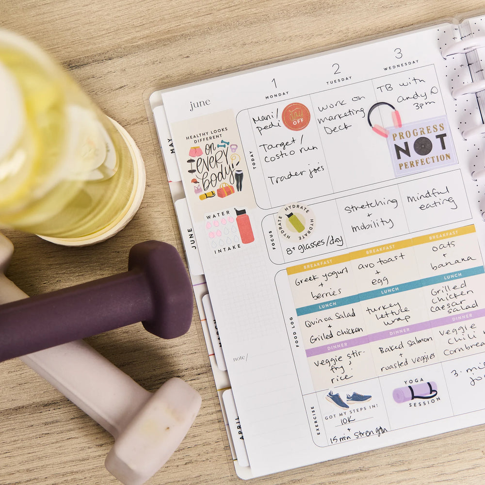 Planner with fitness tracking notes on a wooden surface with dumbbells and a bowl.