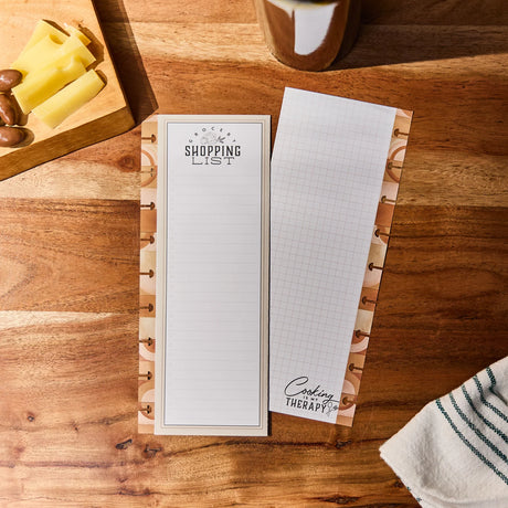 Notebook with a shopping list and cooking-themed pages on a wooden surface.
