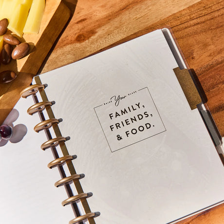 Notebook with 'Family, Friends, & Food' quote on a wooden surface with cheese and nuts.