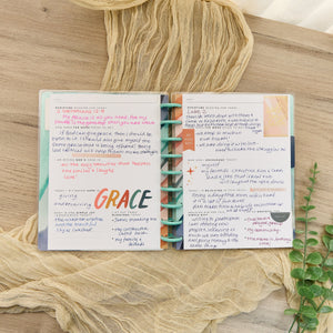 Happy Planner Watercolour Grace Faith Guided Journal with colourful pages on a wooden surface with fabric and plant