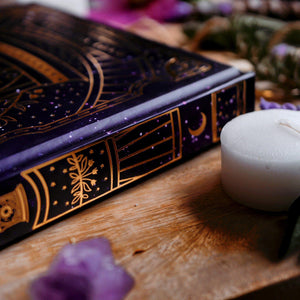 Decorative book with purple cover and gold accents on a wooden surface with a candle and crystals.